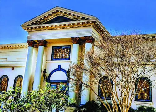 A white building with columns and arch windows, featuring ornamental detailing. A tree with sparse foliage stands in the foreground. Clear blue sky in the background.