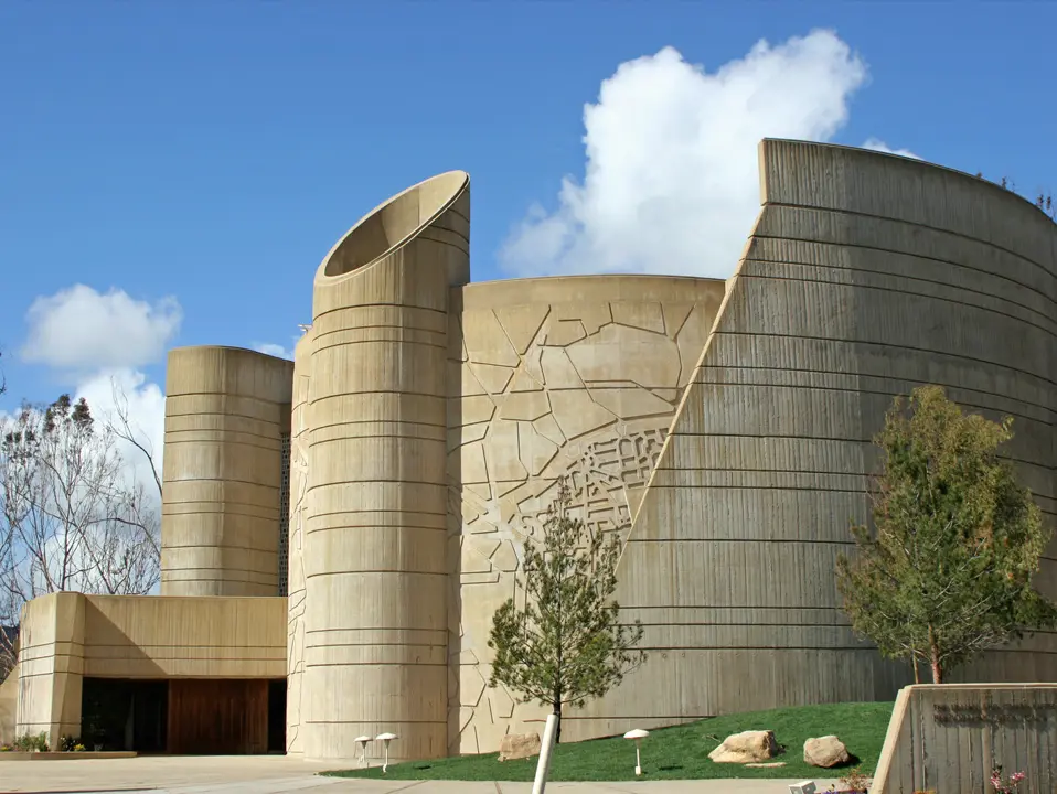 A modern building with curved, textured concrete walls and abstract designs, set against a blue sky with scattered clouds and surrounded by trees.